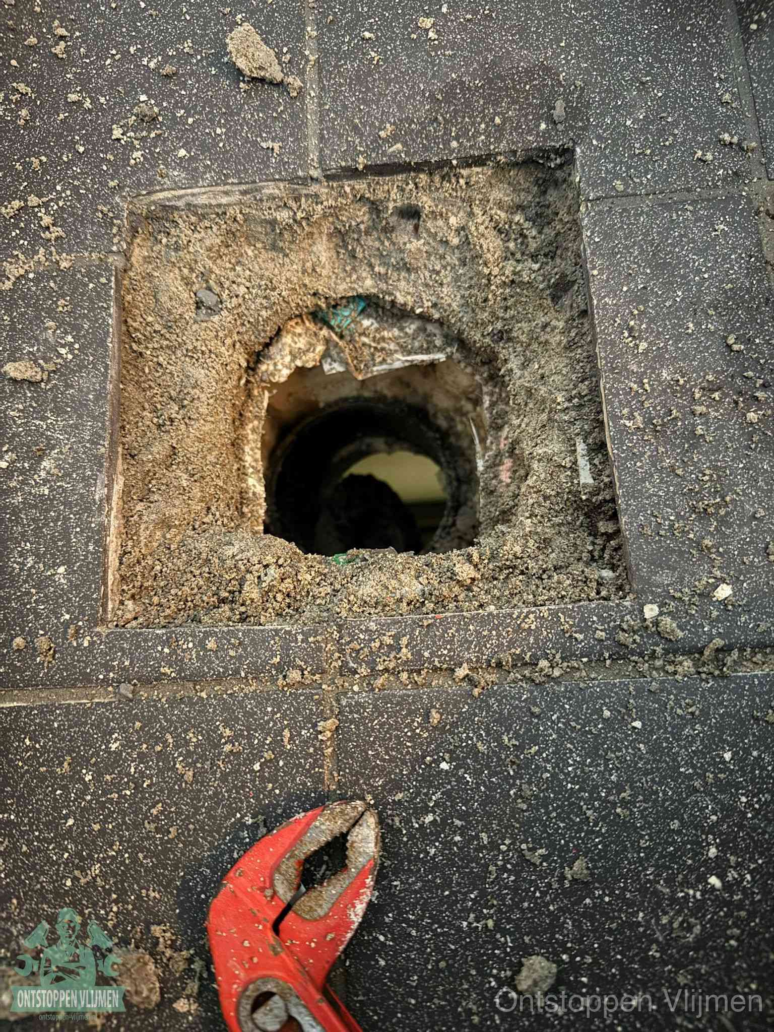 Een geopend rioolputje in een betontegelvloer met losgemaakt rooster en een zichtbare ronde afvoerbuis, het achterhalen waar de afvoerverstopping zit in Vlijmen