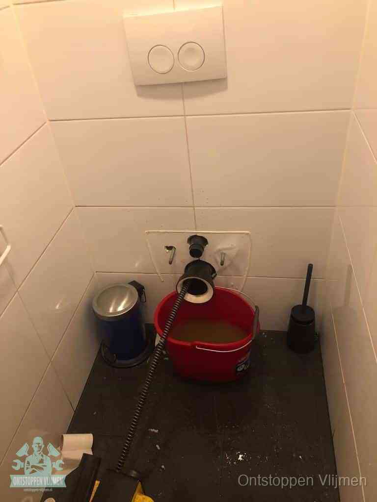 Een ontstopte wand-wc met verwijderde toiletpot waar een ontstoppingsveer in de wanduitgang verdwijnt, het achterhalen waar de verstopping zit in Vlijmen