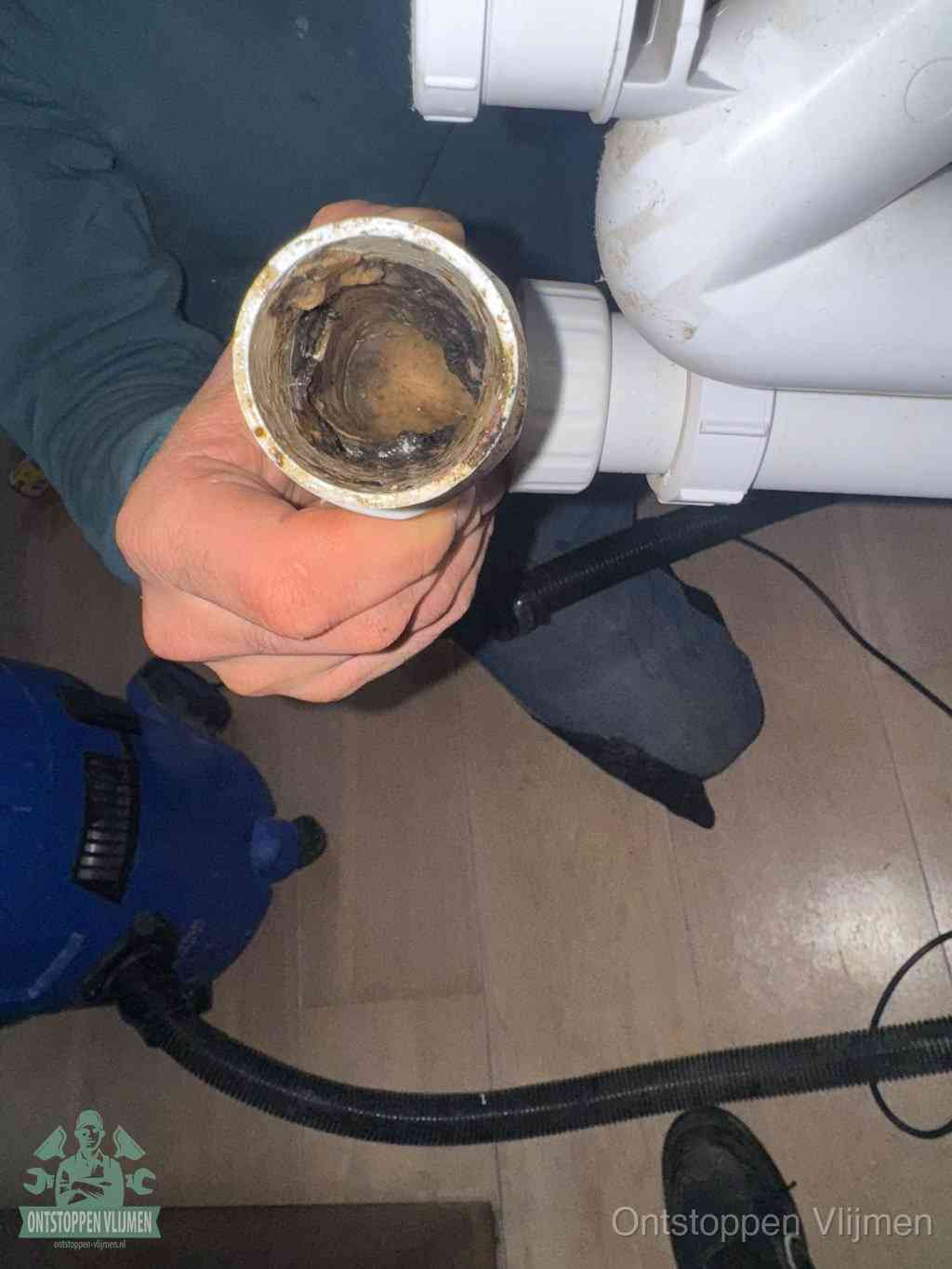 Een hand houdt een losgekoppeld PVC-afvoerbuisstuk vast dat van binnen volledig is dichtgeslibd met vet, vuil en aanslag, de meest voorkomende oorzaak van een verstopte afvoer in Vlijmen
