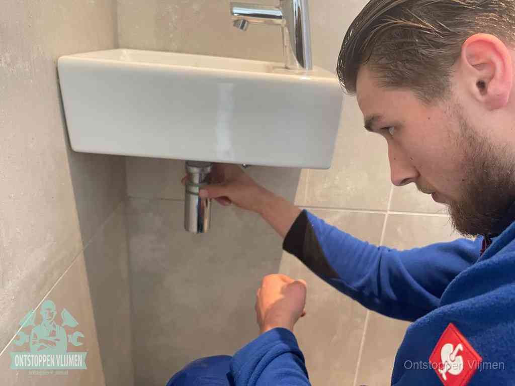 Een monteur draait met de hand de zwanenhalssifon onder een fonteinwasbak in de toiletruimte los om de bron van rioollucht in Vlijmen te vinden