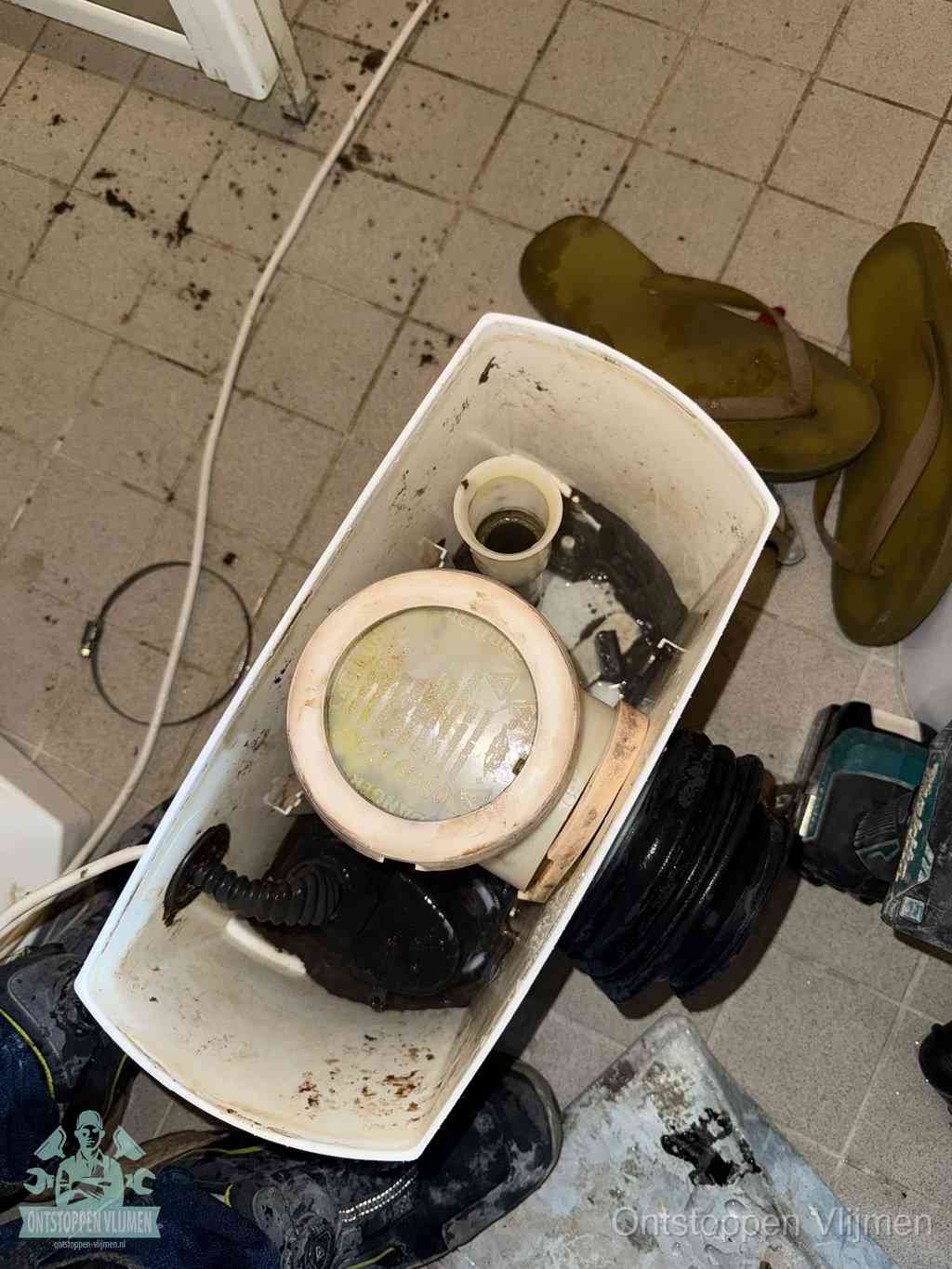 Een gedemonteerde toiletpot op de vloer met ontstoppingsveer en gereedschap ernaast, het verwijderen van een vastzittend voorwerp uit de wc in Vlijmen